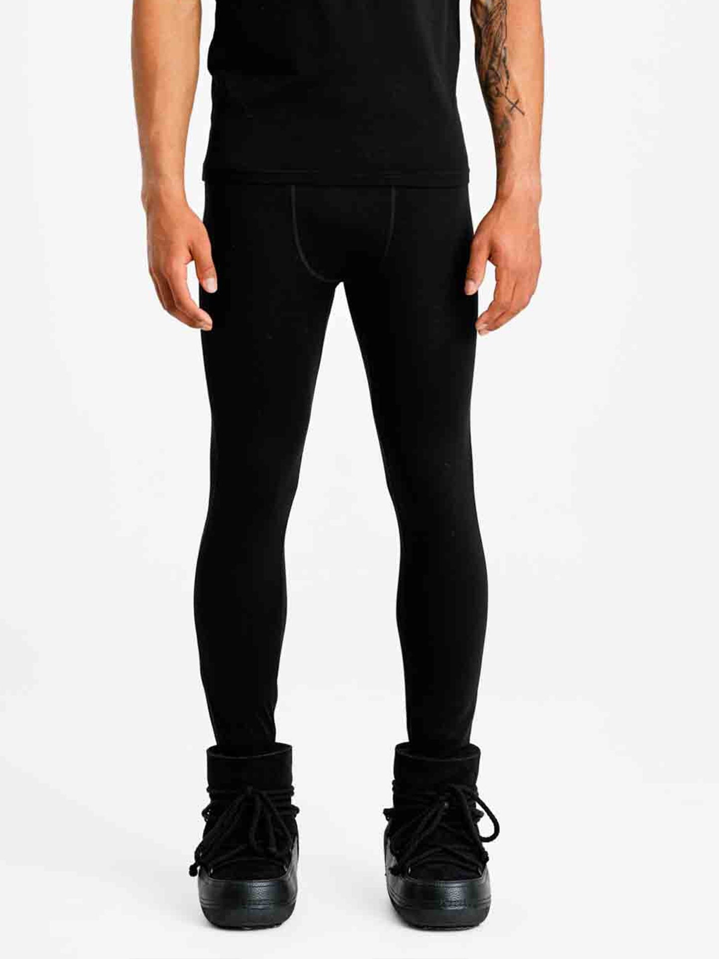 A person stands against a plain white background wearing black leggings, a black shirt, and black high-top shoes. Only the lower half of their body is visible, showcasing the details of "Sno Merino Long Johns Bottoms Men" by We Norwegians under their attire for added breathability and odor resistance.