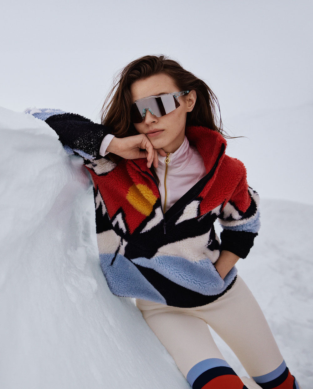 A person in reflective sunglasses and a colorful, oversized Snowfun Sweater Women by We Norwegians leans against a snowy slope. The wintry landscape forms the backdrop, while the person exudes a stylish, adventurous vibe.