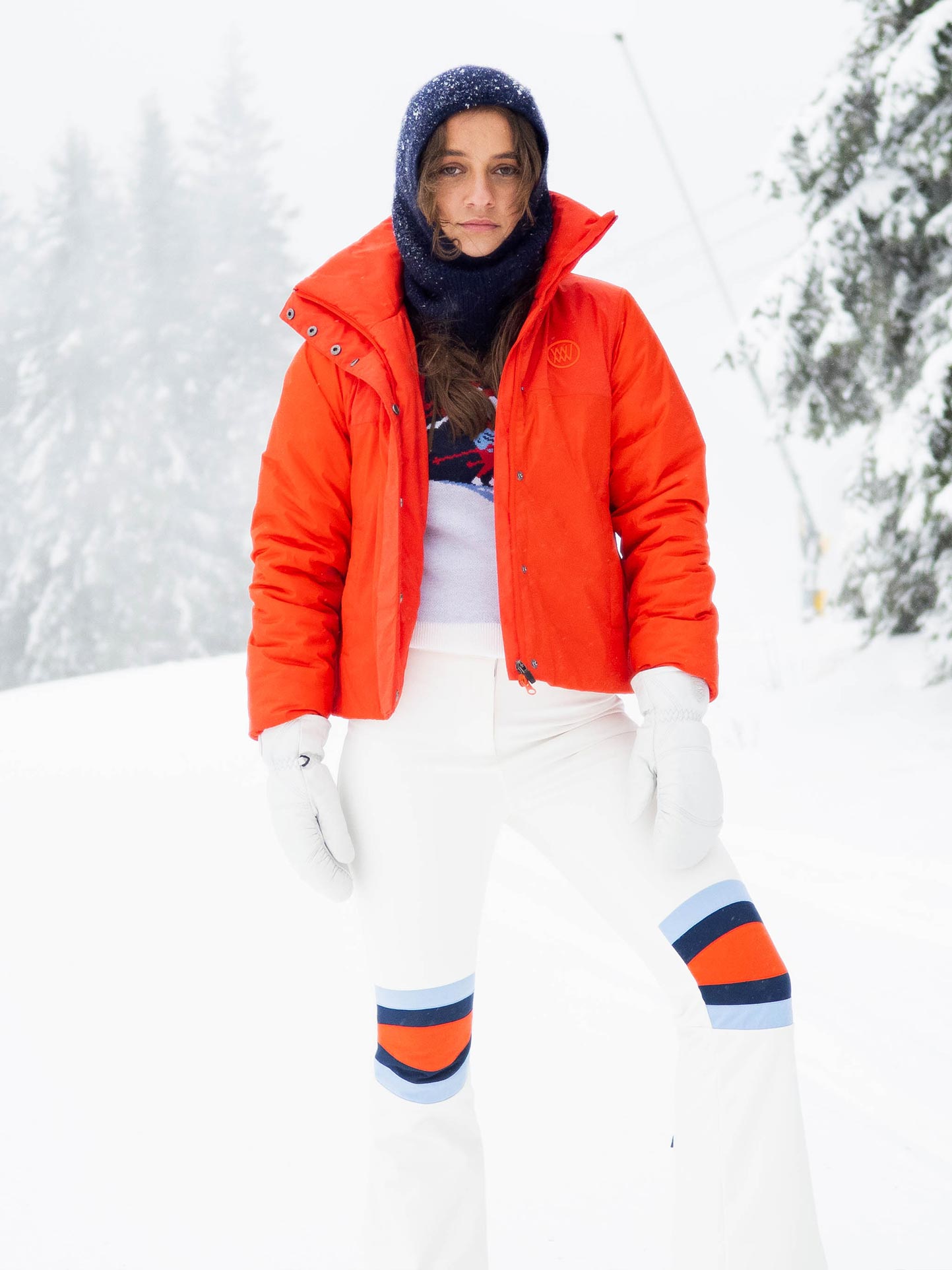 A person stands on a snowy landscape, wearing a bright orange jacket, **We Norwegians Snowfun Ski Pant Women** with blue and orange stripes, white gloves, and a blue head covering. Snow-covered trees and a ski lift are visible in the background. The person looks directly at the camera.