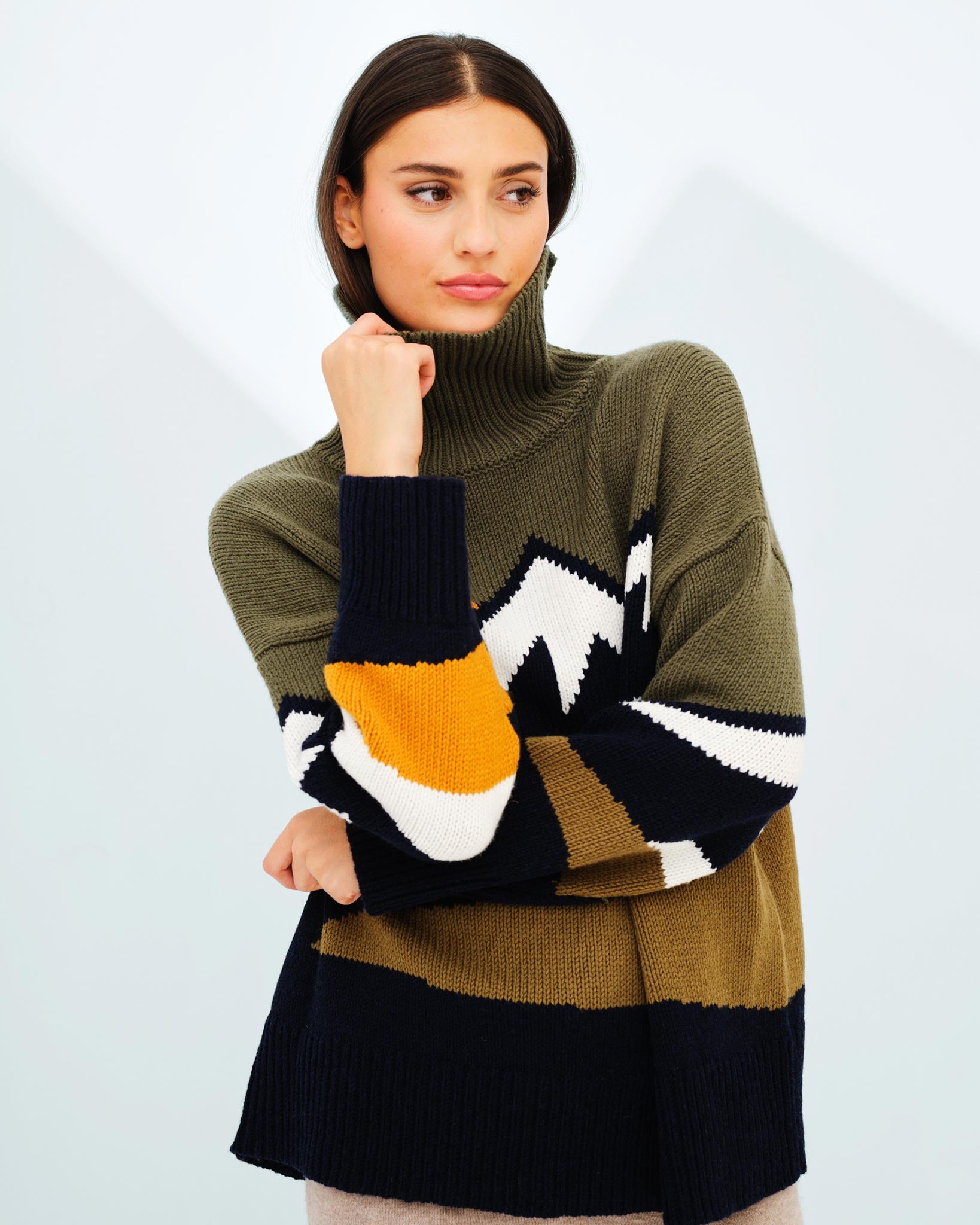 A woman with long, dark hair is wearing a high-neck, oversized Snowmountain Sweater by We Norwegians, featuring an abstract, colorful design. She appears to be thoughtfully posing with her hand near her chin, looking slightly away from the camera. The light and minimalist background perfectly highlights the fall fashion vibes.