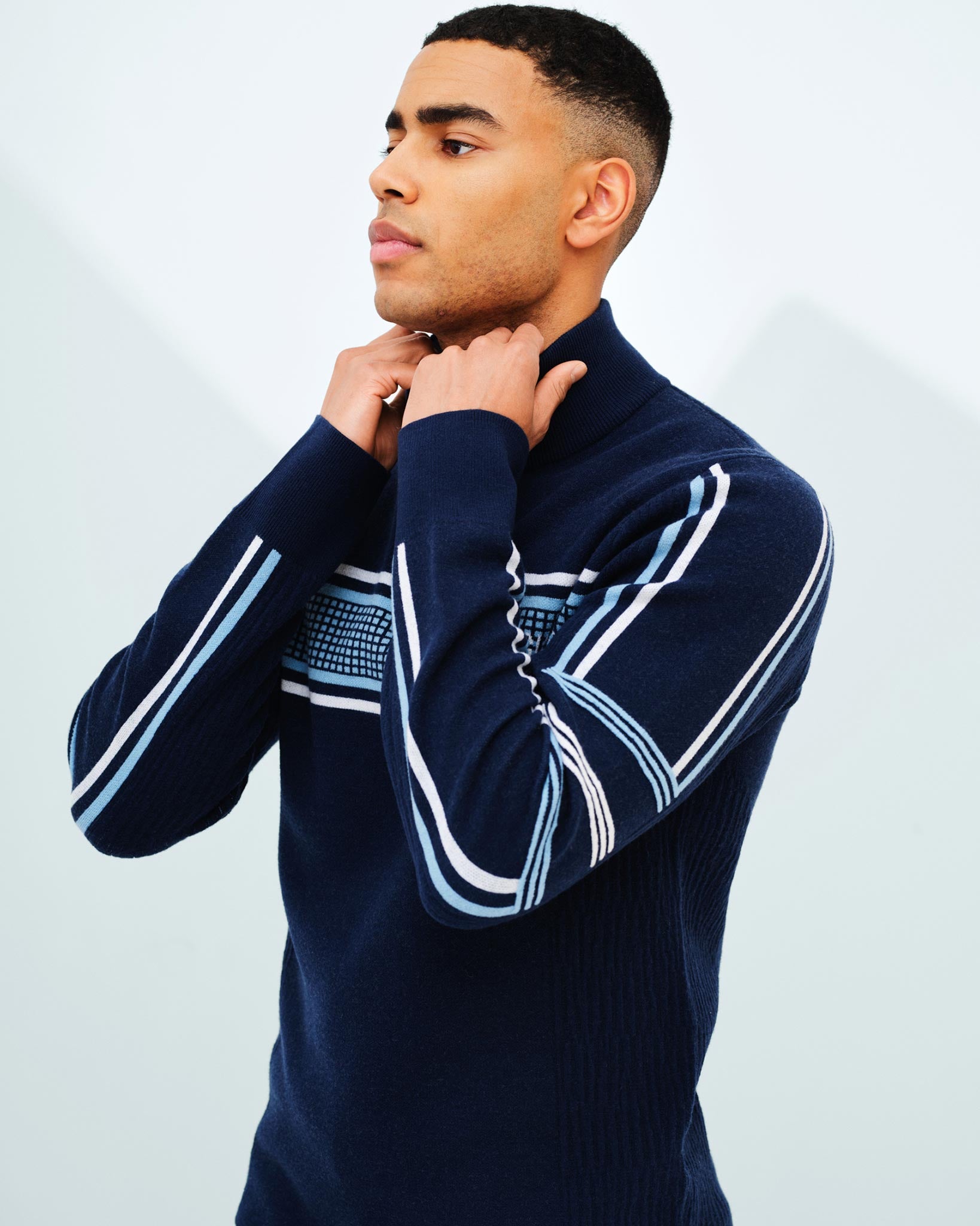 A person with short dark hair is standing against a light background, wearing the Stryn Zipup Men sweater by We Norwegians, made of ultrafine merino wool. The navy blue sweater features white and light blue stripe patterns reminiscent of retro ski designs. They are adjusting the collar with both hands and have a calm, focused expression.