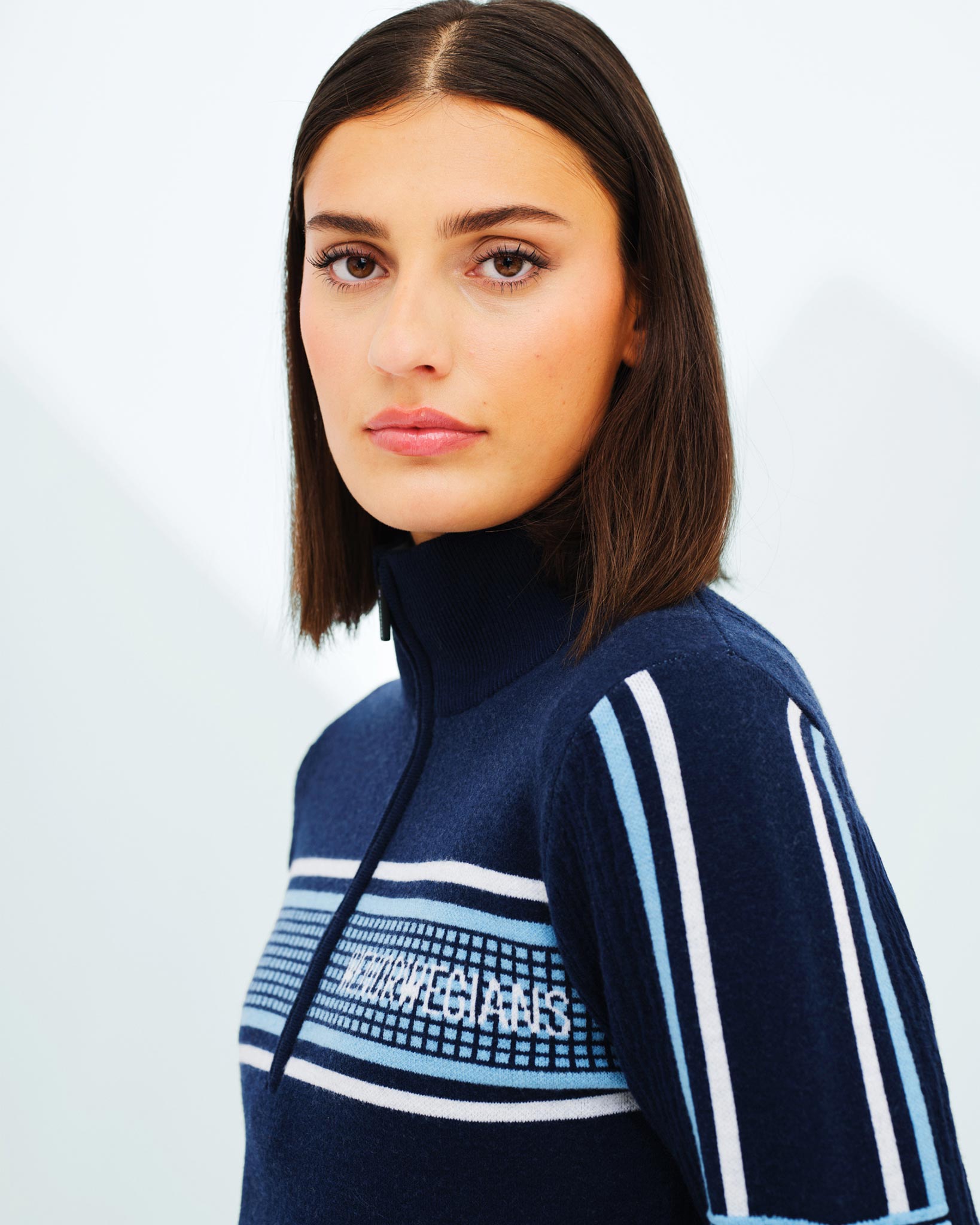 A woman with shoulder-length brown hair is looking at the camera with a neutral expression. She is wearing the "Stryn Zipup Women" sweater by We Norwegians, which features navy blue, white, and light blue horizontal stripes and a zip-up collar, made from ultrafine Merino wool. The background is plain and light-colored.