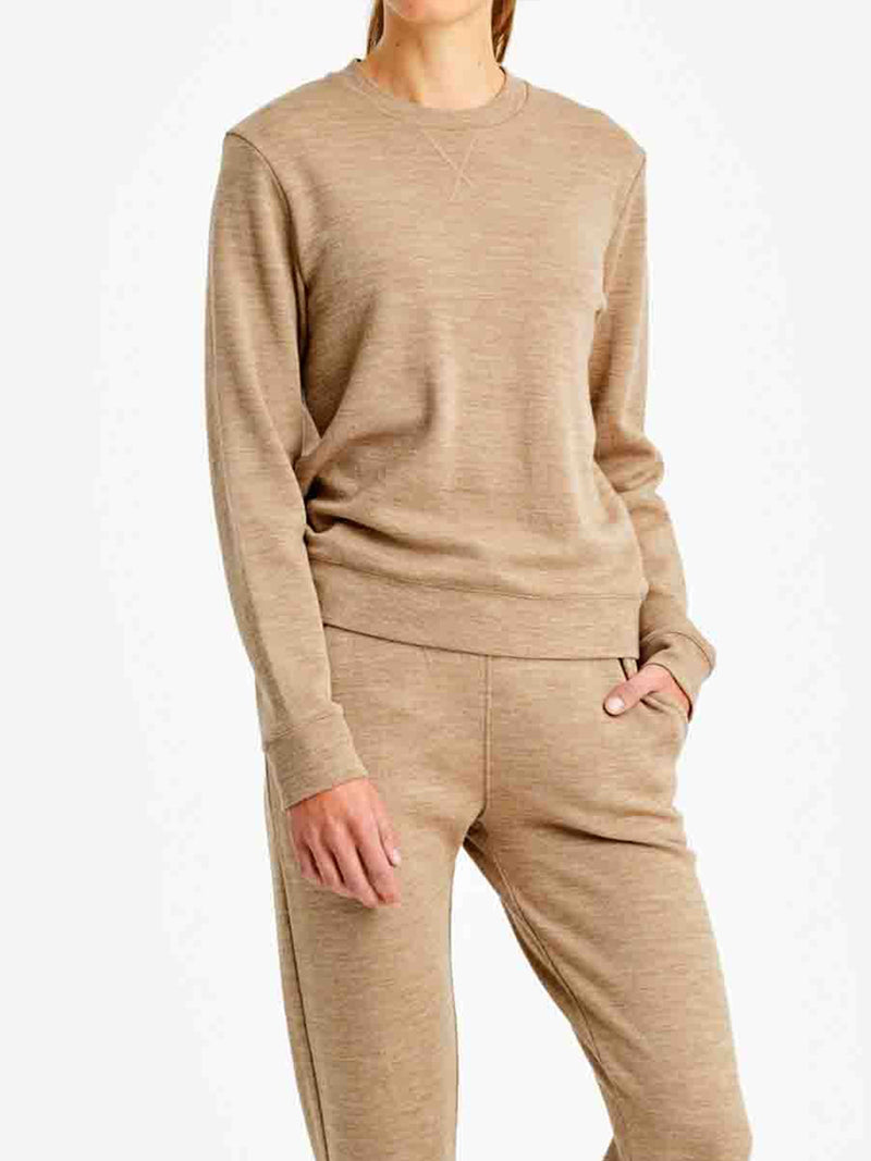 A person is wearing a matching beige long-sleeve top and pants. The top is the Tind Crewneck Women sweater by We Norwegians, featuring ribbed cuffs, and the pants are relaxed-fit joggers with pockets, embodying a luxury sporty style. The person's left hand is in their pocket. The background is plain white.