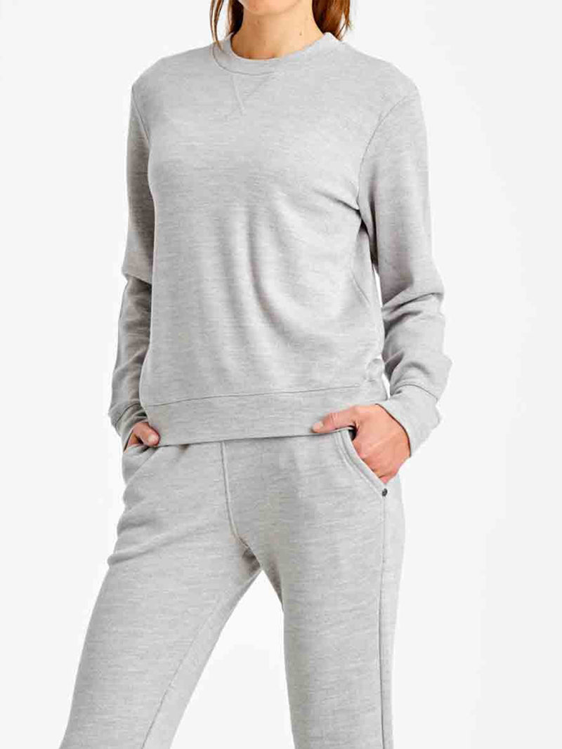 A person stands with hands in their pockets, wearing the We Norwegians Tind Crewneck Women in a casual, light gray hue paired with matching sweatpants. The plain white background accentuates the comfortable and relaxed ensemble. Crafted from luxurious sporty style fabrics, this outfit exudes effortless ease. The individual's face is not visible in the image.