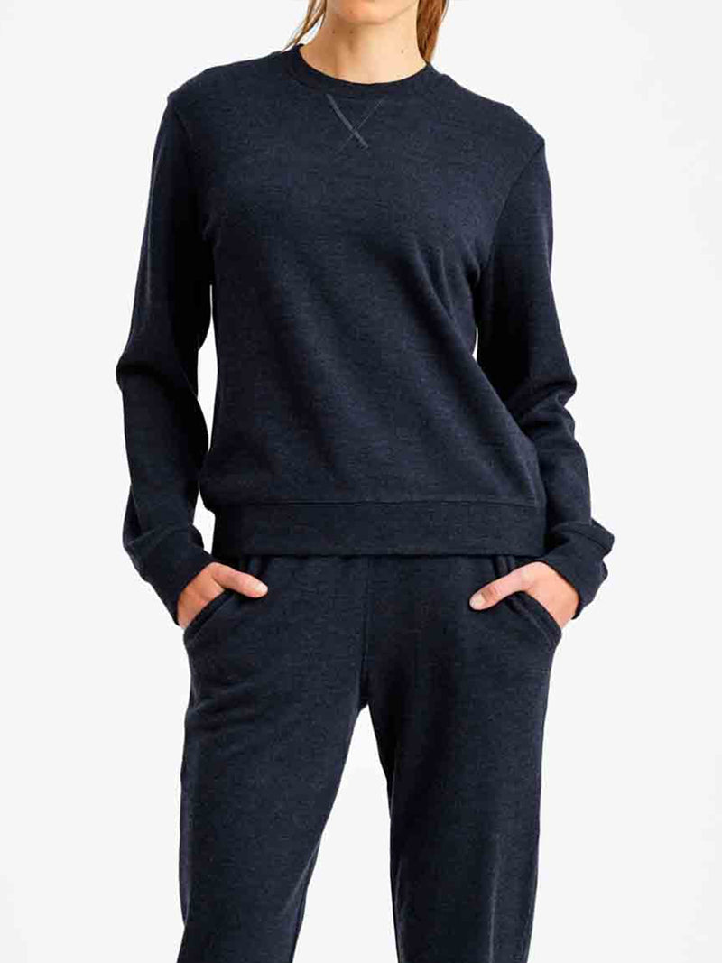 A person wearing a dark-colored, luxurious Tind Crewneck Women sweatshirt from We Norwegians and matching sweatpants stands with their hands in their pockets. The background is plain white. Only the upper body and part of the legs are visible.