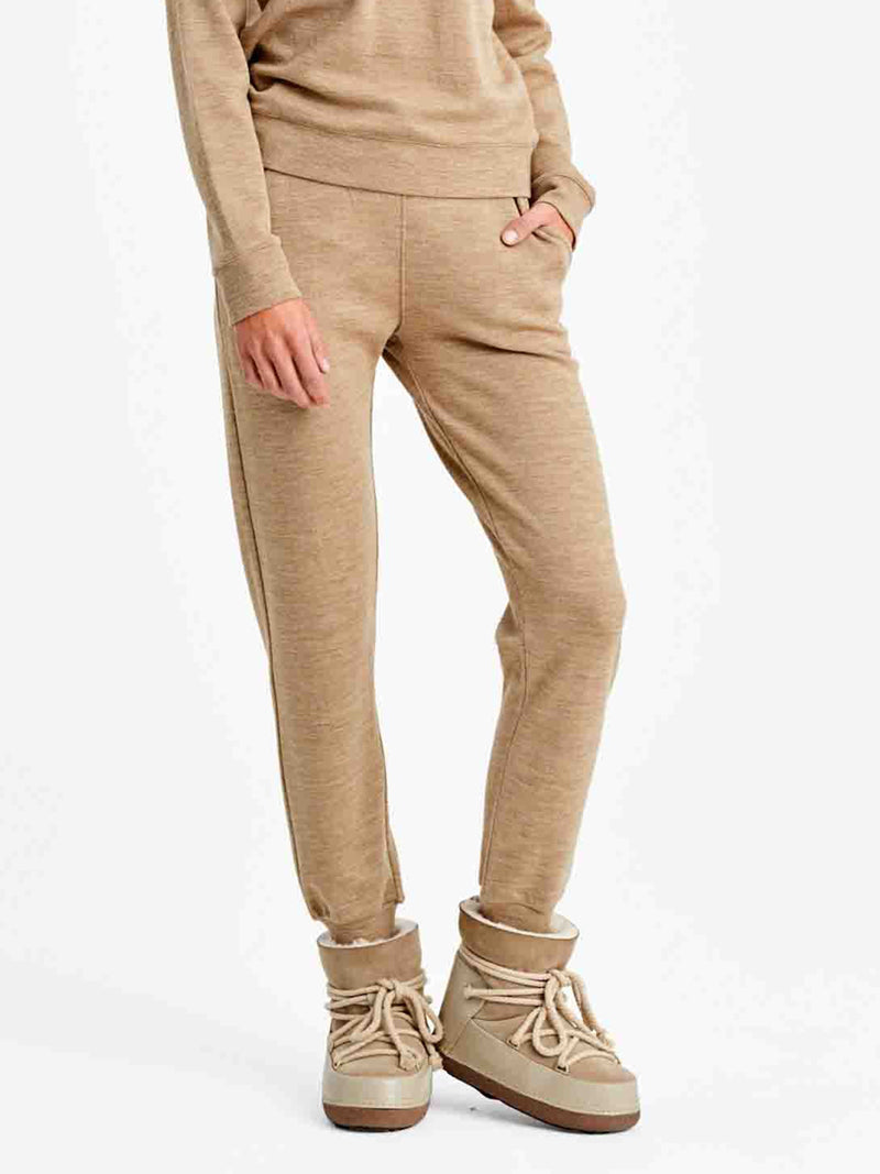 A person stands with one hand in the pocket of their tan Tind Jogger Women pants by We Norwegians. They are wearing matching tan boots with thick laces and a Merino wool sweater, showcasing a relaxed, off-duty style against a plain white background.