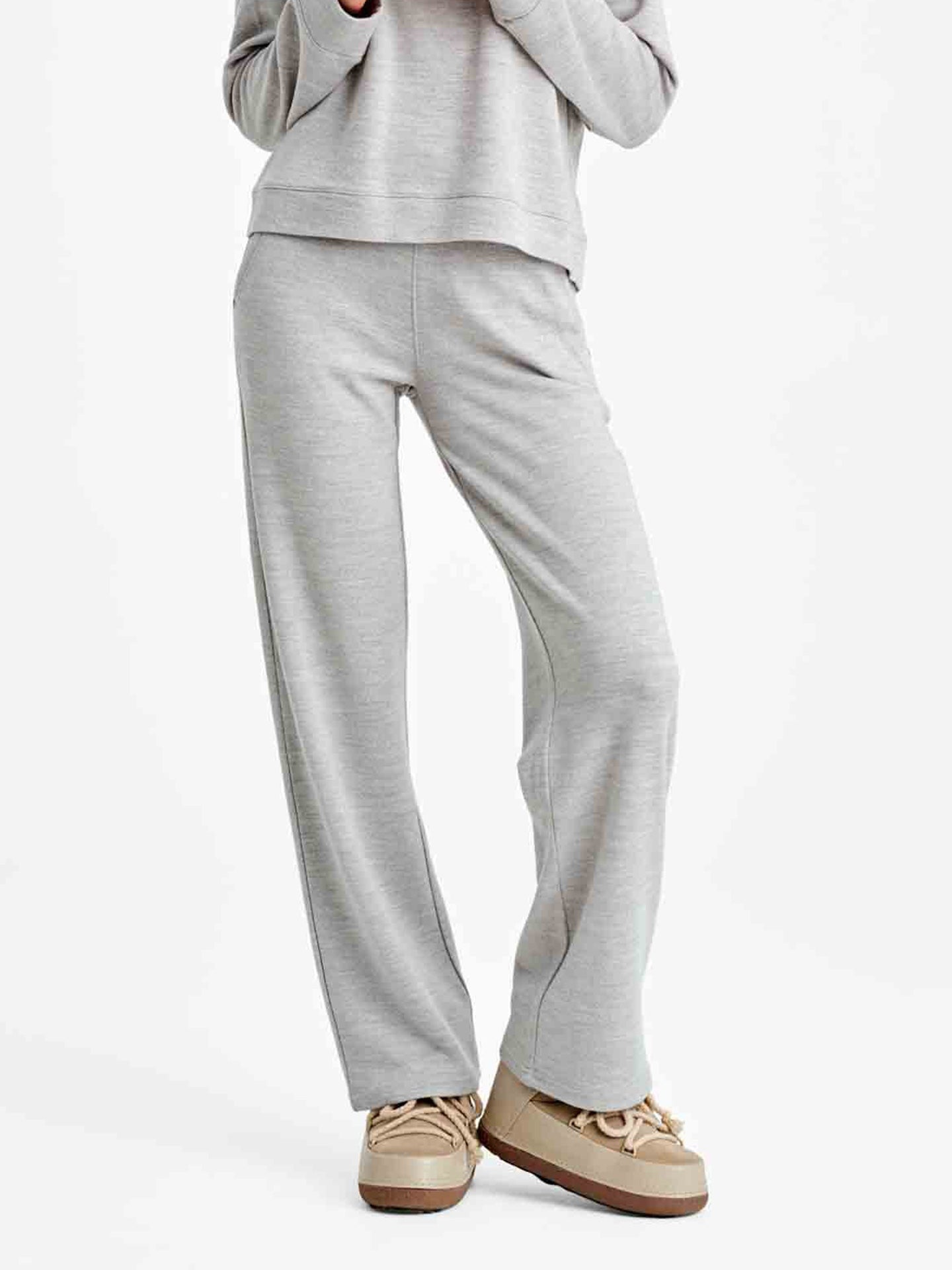 A person is standing against a plain background, wearing the Limited Time Bundle: Tind ZipUp & Tind Straight Leg Pant Women by We Norwegians, which consists of a matching light gray loungewear set made from soft Merino wool. They are also wearing beige lace-up shoes. The person's face is not visible in the image.