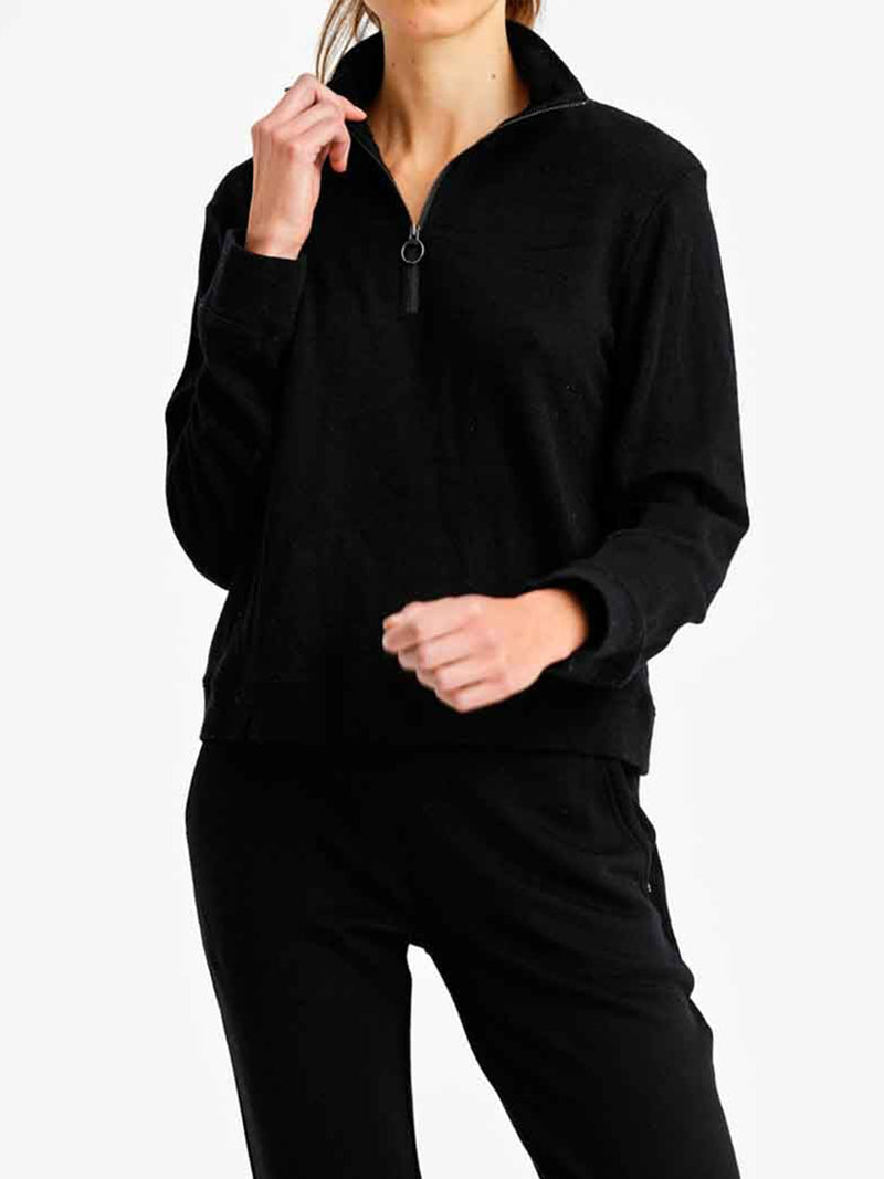 A person is wearing a long-sleeved black We Norwegians Tind Zip Up Sweater from the Limited Time Bundle: Tind Zip Up & Tind Jogger Women, made from Merino wool, with the zipper partially undone, as well as black pants. The person is standing against a plain white background, with their right hand touching their face and their left hand positioned mid-air.