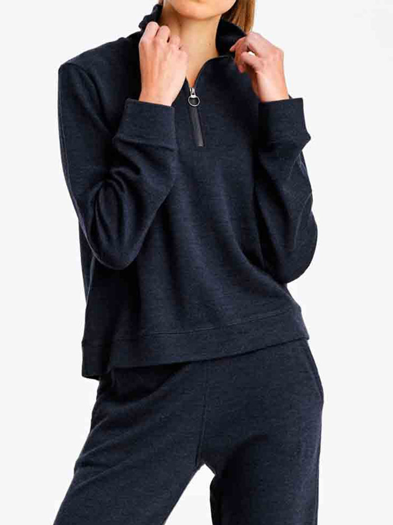 A person dressed in the Limited Time Bundle: Tind Zip Up & Tind Jogger Women from We Norwegians is posing with hands adjusting the zipper on the stylish, half-zip Tind Zip Up Sweater. The comfortable and casual Merino wool outfit stands out against the plain background.