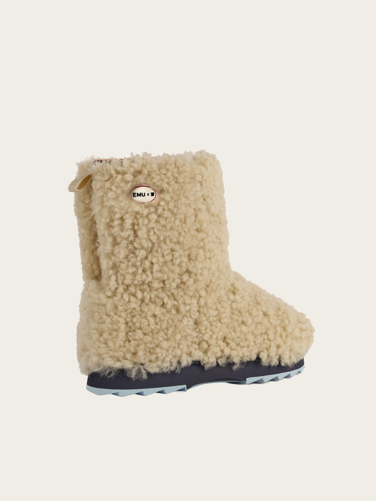 A single fuzzy beige boot crafted from teddy sheepskin, featuring a curly wool texture, a dark blue sole, and a small “EMU” tag on the side, is displayed against a plain off-white background. The EMU x WN Sharky Lo Teddy by We Norwegians showcases premium Australian sheepskin for ultimate comfort and style.