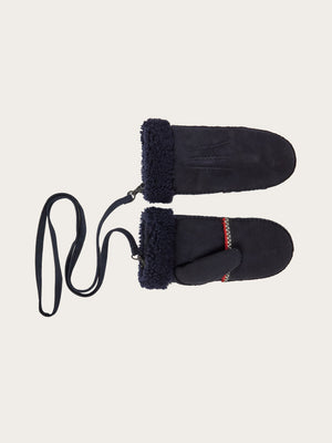 The EMU x WN Montana Mittens by We Norwegians are a cozy cold-weather essential, crafted in dark blue with fuzzy cuffs and joined by a long strap. One mitten is accented with a decorative stitched band in red and white near the wrist, showcasing classic alpine style. Shown here against a plain light background.