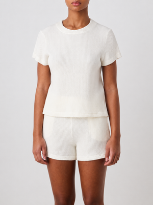 A person stands facing forward, wearing the We Norwegians Waffle Tee Women in a matching off-white set, against a plain light background.