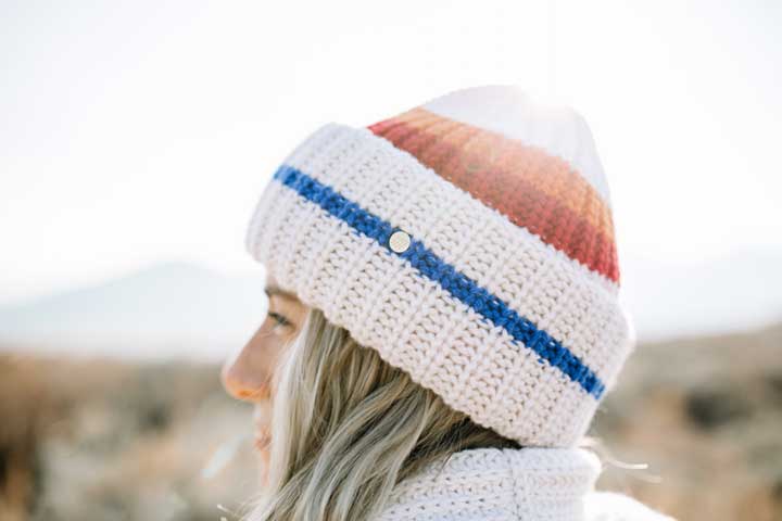 Close-up of a person with long, light-colored hair wearing an Après Ski Hat Women by We Norwegians, featuring red, white, and blue stripes. The background is a blurred outdoor setting with mountains. Sunlight softly shines on the 70's ski fashion beanie.