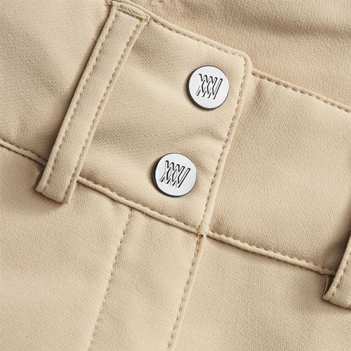Close-up of sand-colored, high-waisted Lista Shorts for women from We Norwegians, focusing on two white buttons adorned with a stylized "XX." The waistband and belt loops exhibit detailed stitching, showcasing the refined Swiss Schoeller fabric. The material's texture is slightly textured and neatly finished.