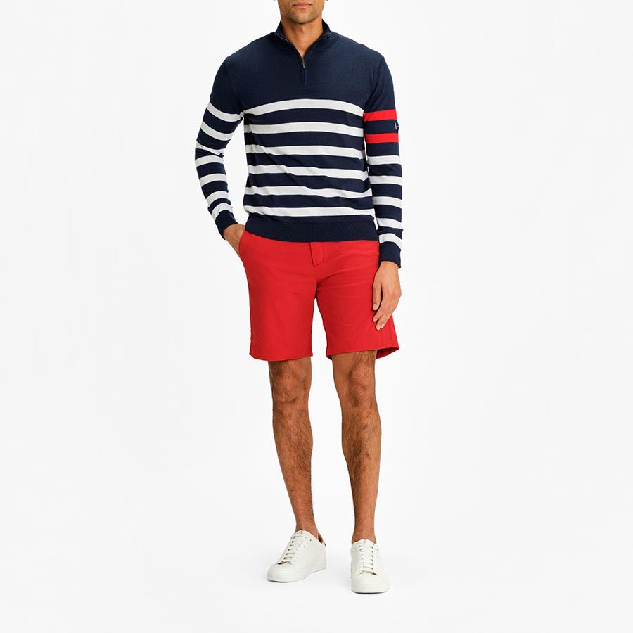 A person is wearing a navy and white striped sweater with a red stripe on one arm, paired with the Lista Shorts Men Red from We Norwegians. These stylish red shorts are made from Swiss Schoeller fabric for 4-way stretch comfort. The look is completed with white sneakers against a plain white background, perfectly embodying the sleek style of the Lista product line.