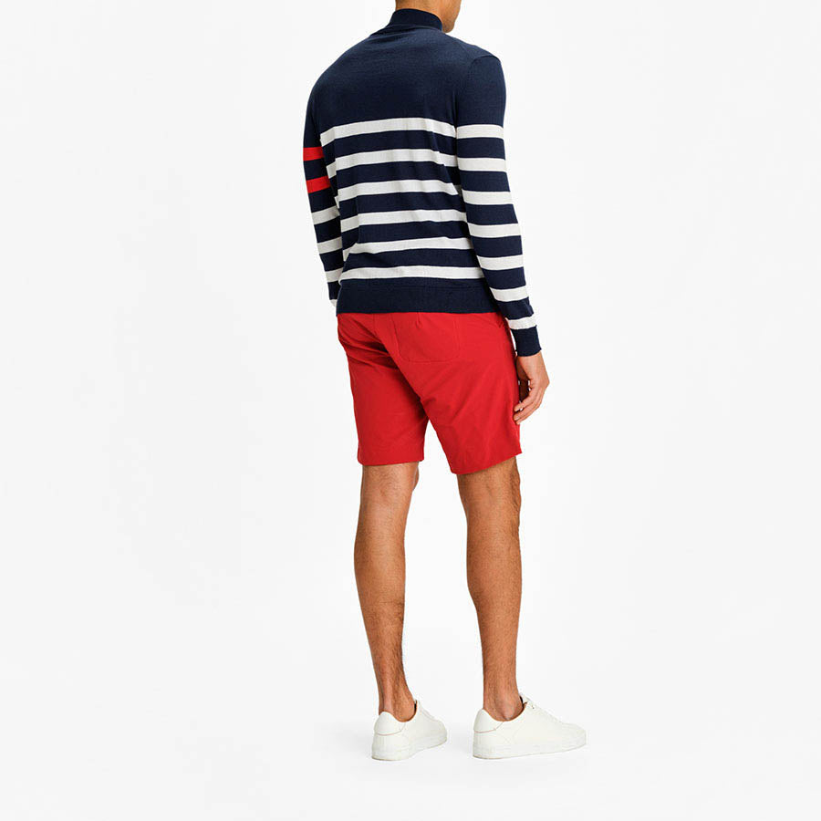 A person stands facing away, wearing the We Norwegians Hjellestad Zip Up Men Navy sweater, which features a red stripe on the left sleeve. Paired with red shorts and white sneakers, this versatile garment is perfect for any occasion. The background is plain white.