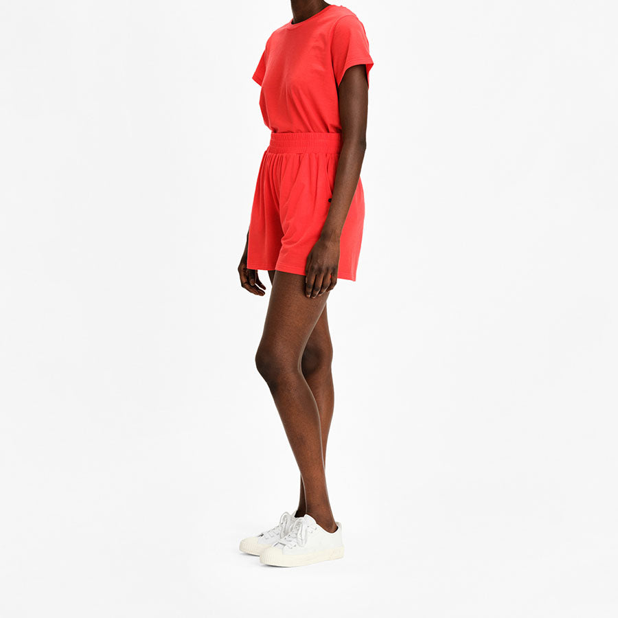 A person is standing against a plain white background, wearing the Salt Classic Top Women Coral by We Norwegians, paired with matching bright red shorts made from breathable Creora-Eco-Soft. They are also wearing white sneakers. The person is facing slightly to the side, with their right arm relaxed by their side.