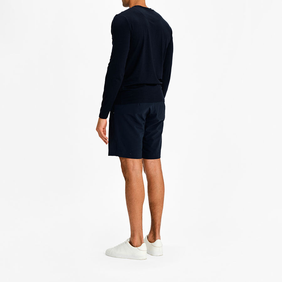 A person in a navy "Salt Longsleeve Men" shirt by We Norwegians and navy shorts is standing with their back facing the camera. They are wearing white sneakers. The background is plain and white.