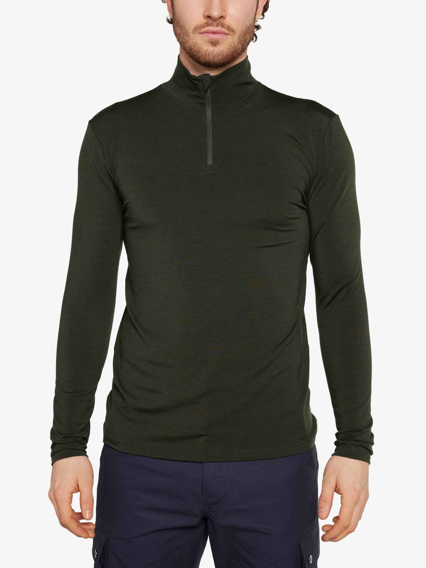 A person stands against a white background, wearing the We Norwegians' Hygge Zip Up Men in Olive Green with dark pants. This Merino wool base-layer top features a high collar, and their hands rest relaxed by their sides.