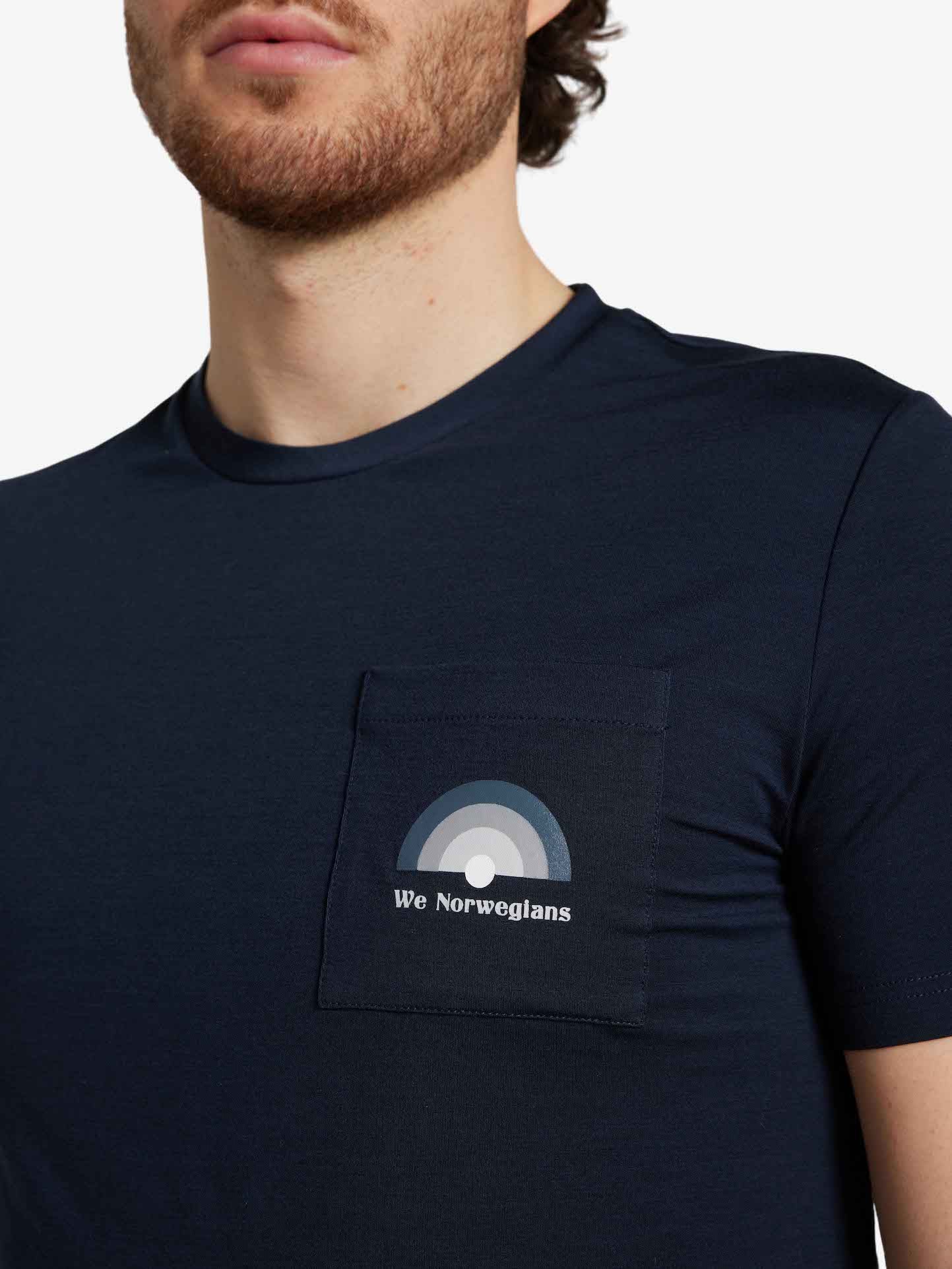 A person wearing the Rainbow Tee Men Navy Blue by We Norwegians, which features a small left chest pocket with a gray-shaded rainbow design and text reading "We Norwegians." Ideal for Spring/Summer, the individual has a partial beard, light skin, and dark hair visible.
