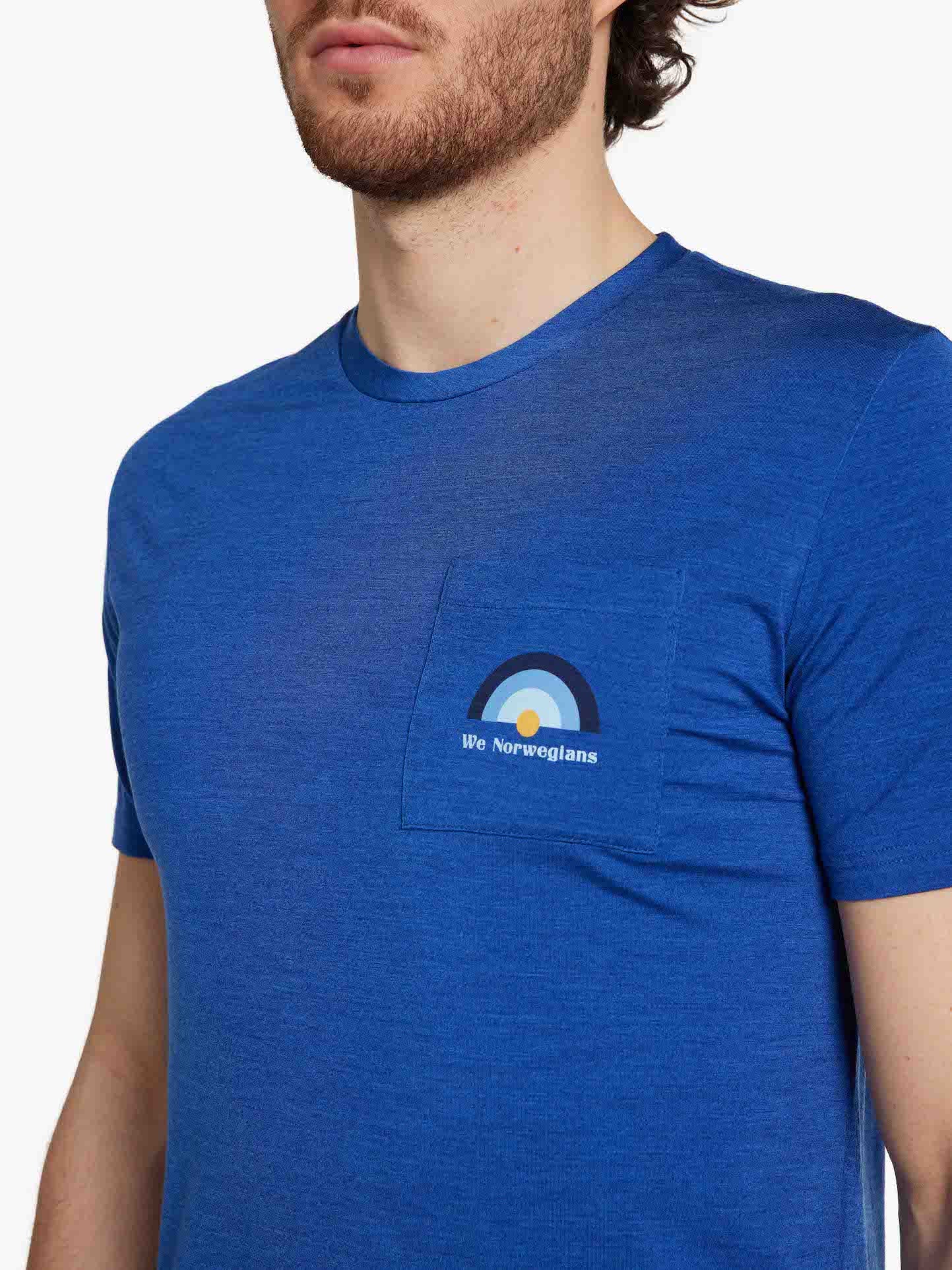 A person wearing the Rainbow Tee Men Royal Blue from We Norwegians, featuring a small left chest pocket with a minimalist semi-circle design and the text "We Norwegians" embedded within it. Ideal for spring/summer, this stylish piece effortlessly combines comfort and elegance against a crisp white background.