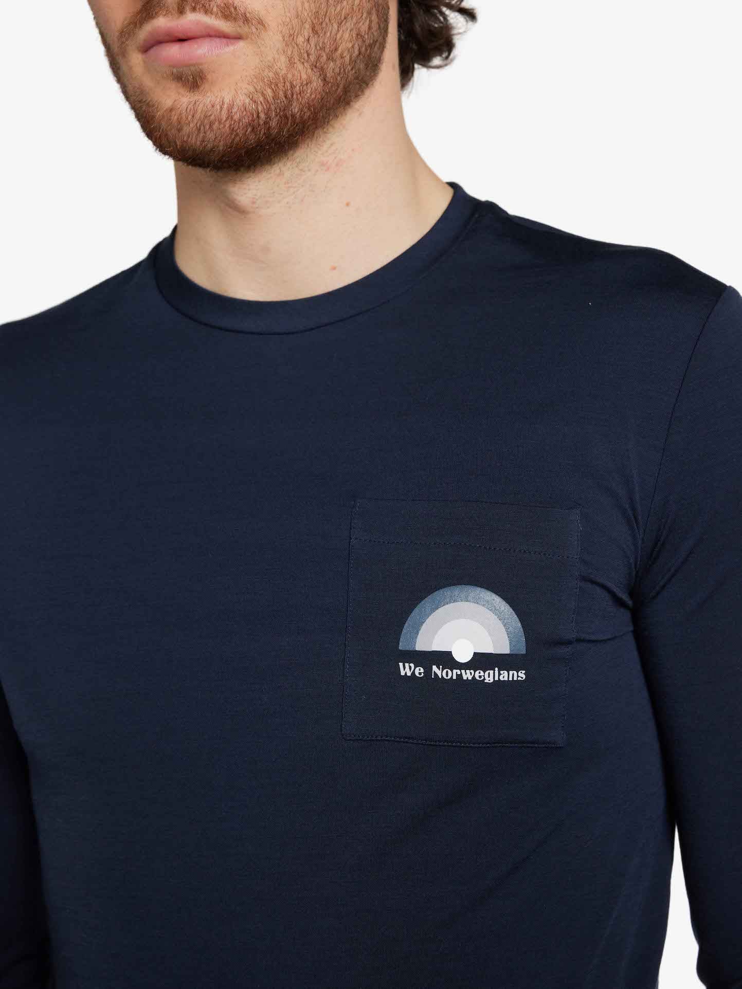 A man with a short beard and wavy hair wears the Rainbow LongSleeve Men Navy Blue by We Norwegians. The shirt has a small front pocket featuring a logo design with overlapping circles and the text "We Norwegians." The man's face is partially out of the frame.