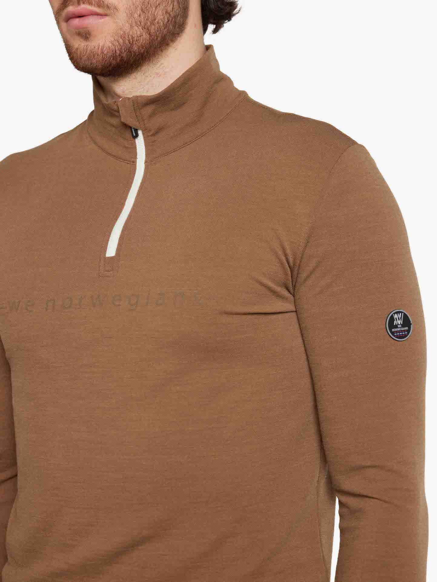 A man with a short beard wearing the Lindesnes ZipUp Men Cinnamon, a brown long-sleeve, half-zip pullover made of Super Fine Merino by We Norwegians. The pullover features a small circular black and white logo patch on the upper left sleeve and the partially visible text "We Norwegians" on the chest. This stylish mid-layer blends perfectly with the white background.