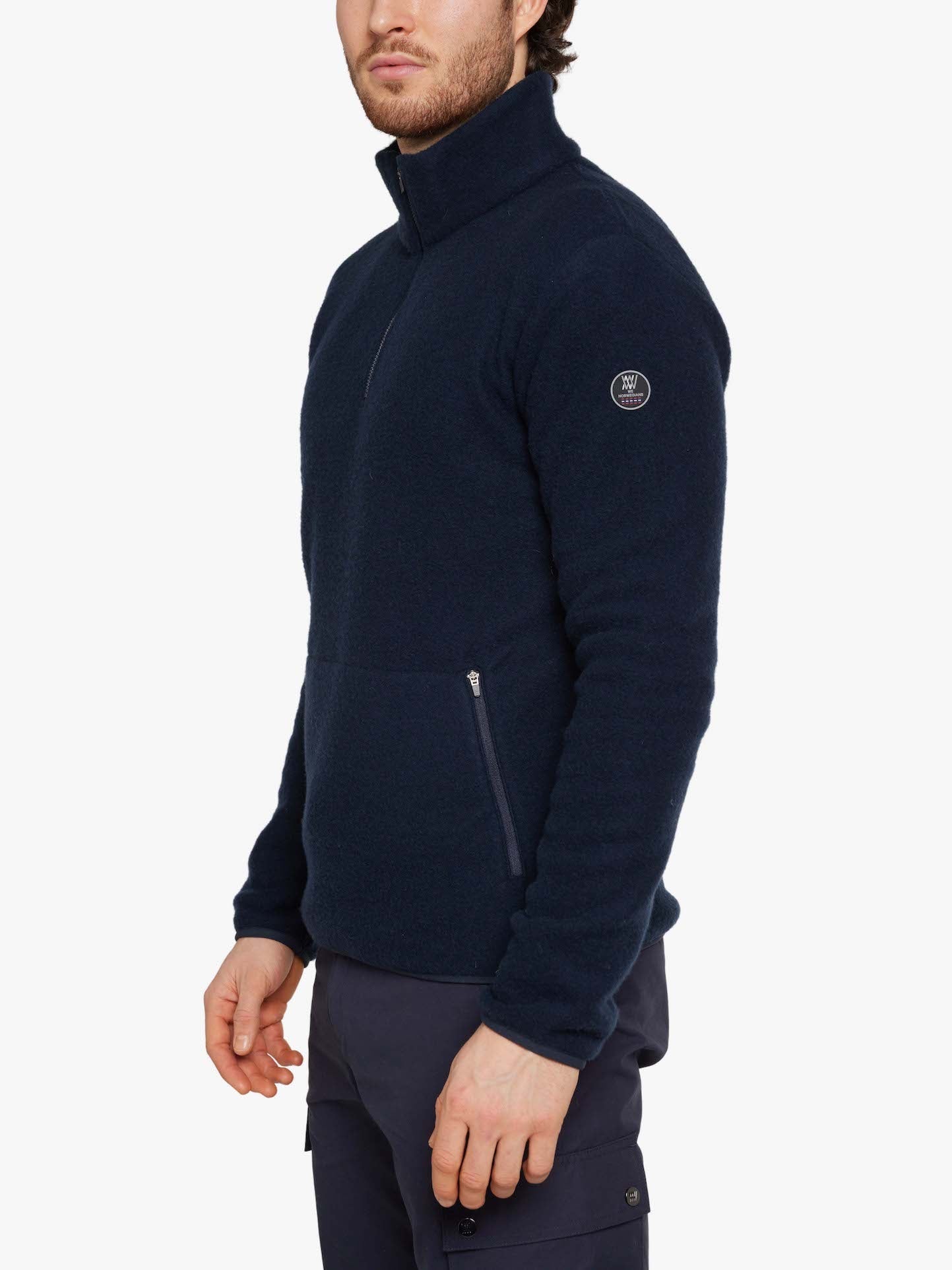 A person is standing sideways wearing a We Norwegians Svalbard Sweater Men in Navy Blue, which features a half-zip front and a logo on the upper sleeve. They are also wearing dark blue pants with a side pocket. The background is white.