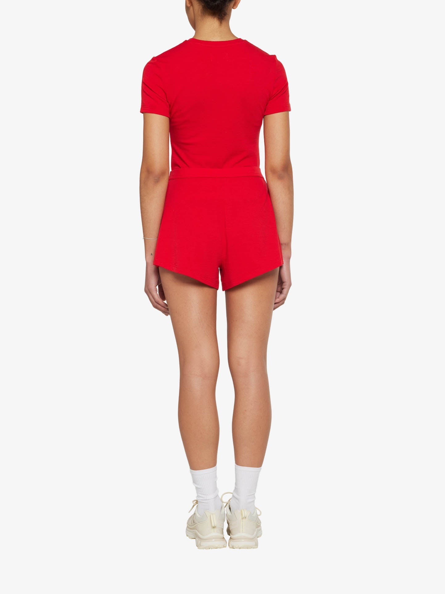 A person is standing with their back to the camera, dressed in a vibrant red matching set featuring a short-sleeved top and We Norwegians Lindesnes Running Shorts Women Red. They are also wearing white socks and white sneakers, against a plain white background.