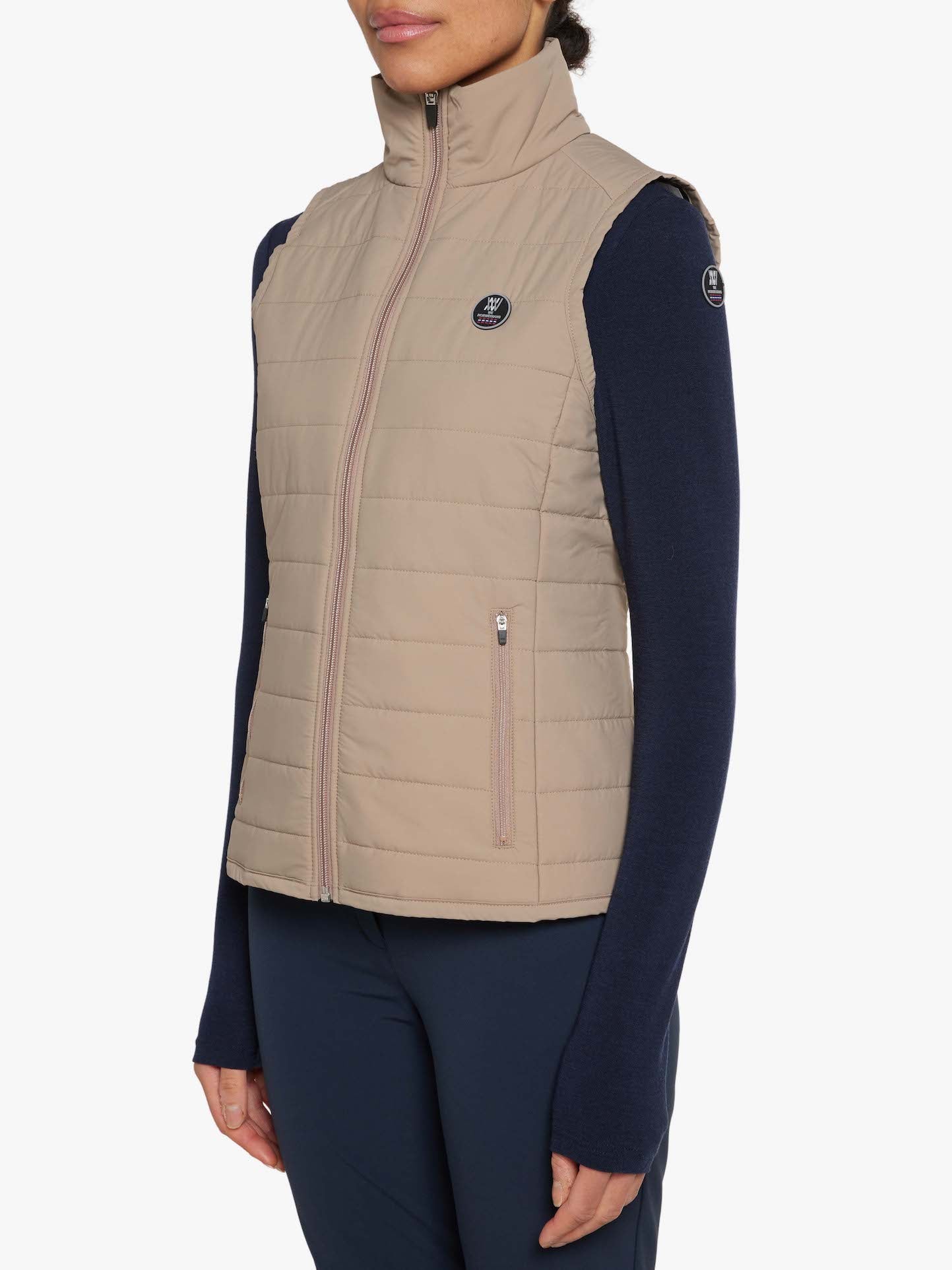 A person is wearing a sand-colored, quilted, sleeveless Lyderhorn vest from We Norwegians over a navy long-sleeve top. The water repellent polyester vest features a high collar, two front zippered pockets, and small round logos on the upper left chest and right shoulder. The person's lower body is not fully visible.